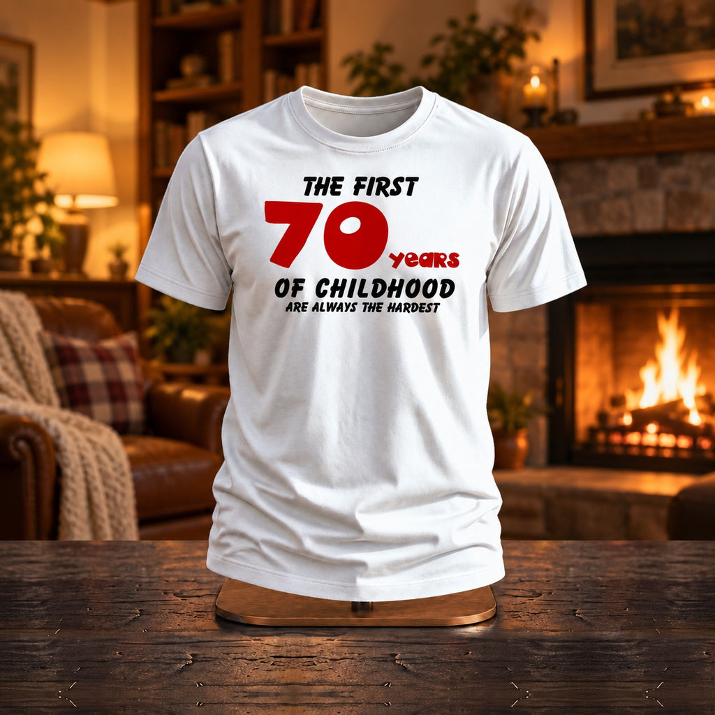 The First 70 Years of Childhood T-shirt in white with a red 70 on a wooden stand by a cosy fireplace - funny 70th birthday gift