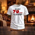 The First 70 Years of Childhood T-shirt in white with a red 70 on a wooden stand by a cosy fireplace - funny 70th birthday gift