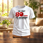 The First 80 Years of Childhood T-shirt in white with a red 80 on a wooden stand in a bright living room - funny 80th birthday gift
