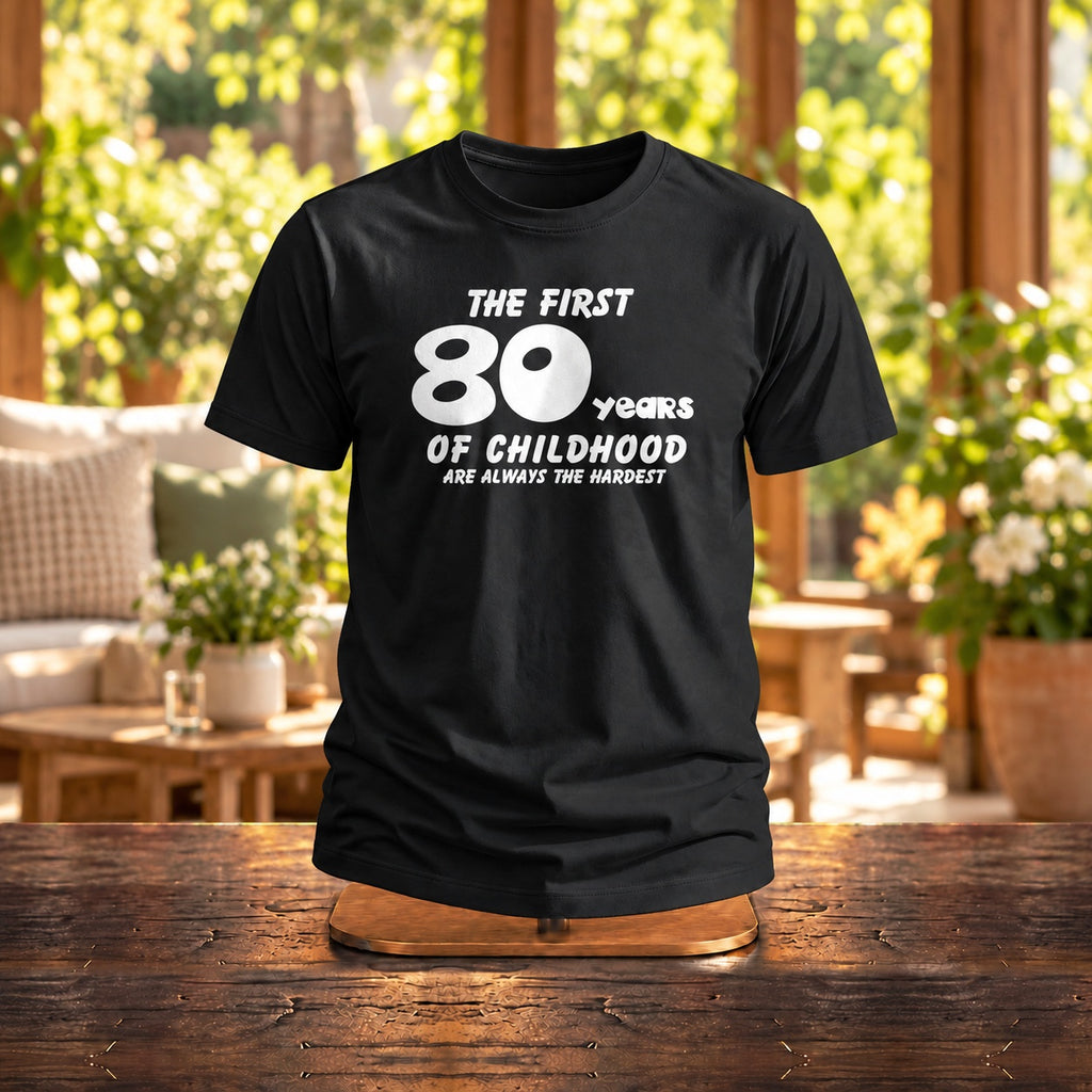 The First 80 Years of Childhood T-shirt in black on a wooden stand in a cosy café with plants - funny 80th birthday gift