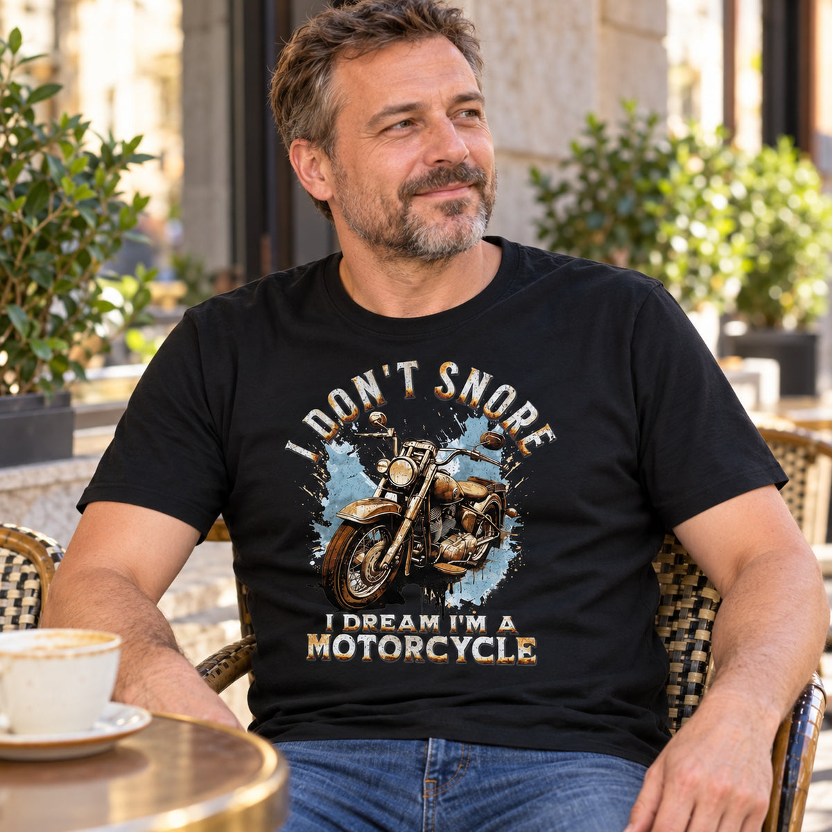 Biker wearing a black I Don't Snore, I Dream I'm a Motorcycle t-shirt at a sunny café table