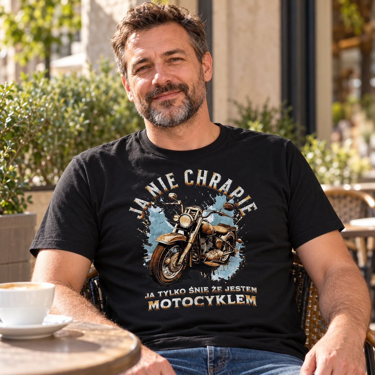 Mężczyzna w czarnej koszulce „Ja Nie Chrapię, Ja Tylko Śnię, Że Jestem Motocyklem” przy stoliku w słonecznej kawiarni