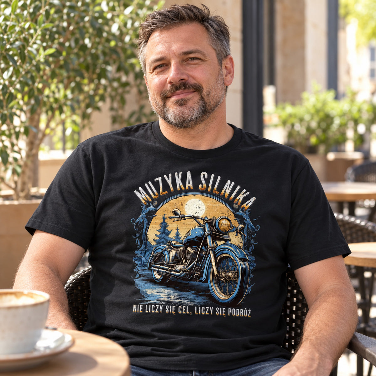 Mężczyzna w czarnej koszulce „Muzyka Silnika” z motocyklem przy stoliku w słonecznej kawiarni