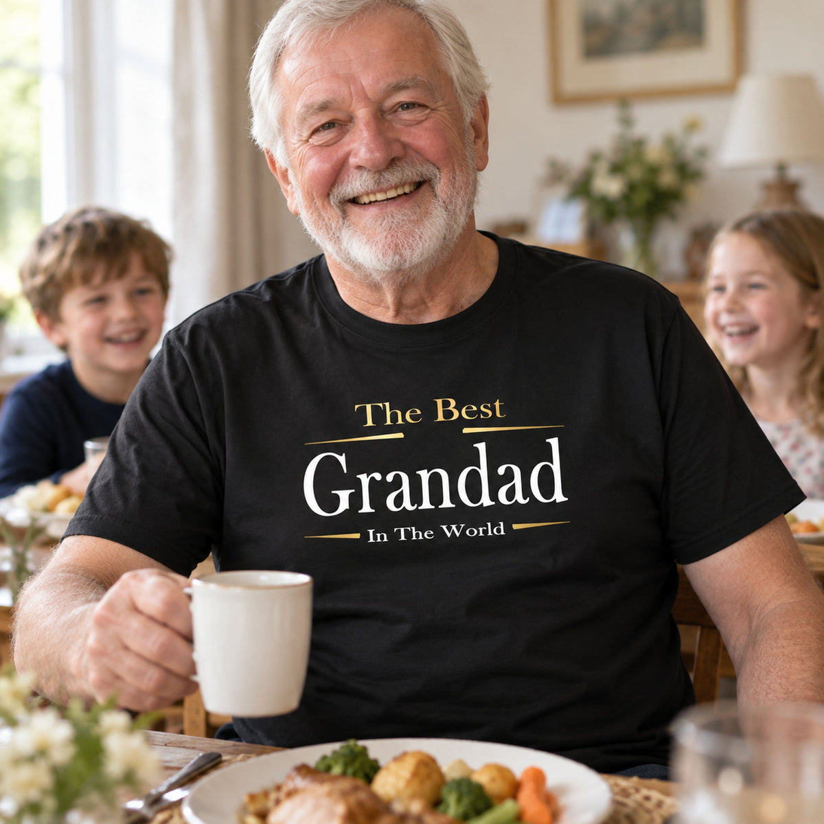 Grandad in a black The Best Grandad In The World Exclusive t-shirt with gold lettering, laughing over a cup of tea at Sunday lunch with two grandkids