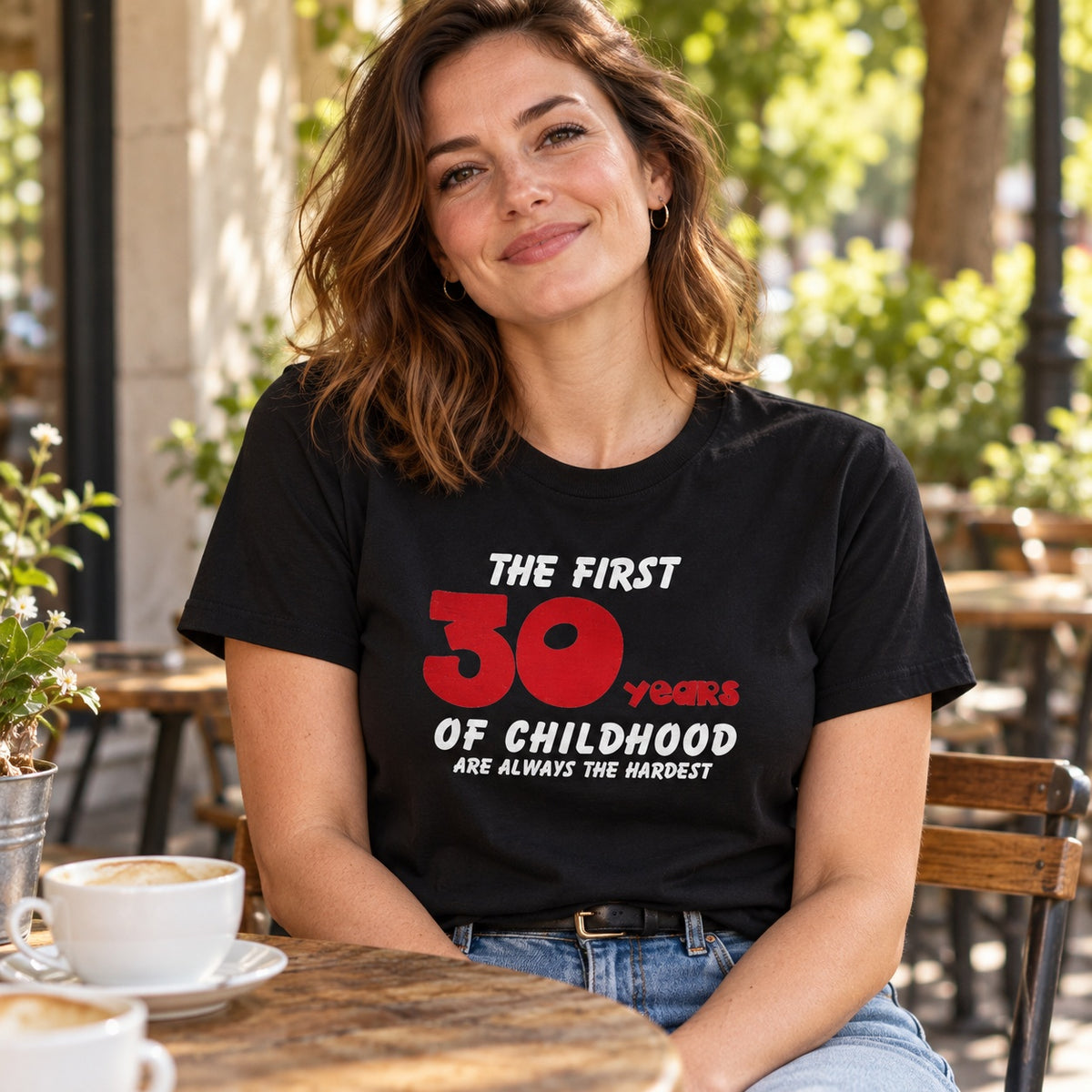 The First 30 Years of Childhood T-shirt in black with a red 30 - smiling woman at a café table with a cup of coffee, 30th birthday gift