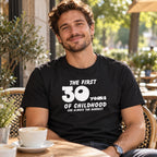 The First 30 Years of Childhood T-shirt in black - smiling man at a café table with a cup of coffee, 30th birthday gift