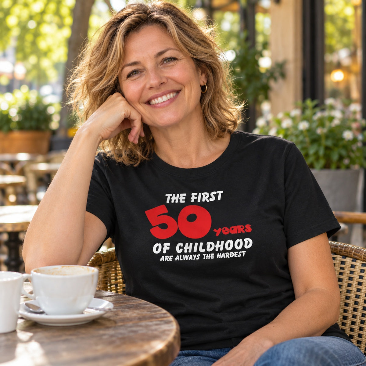 The First 50 Years of Childhood T-shirt in black with a red 50 - smiling woman at a café table with coffee, 50th birthday gift