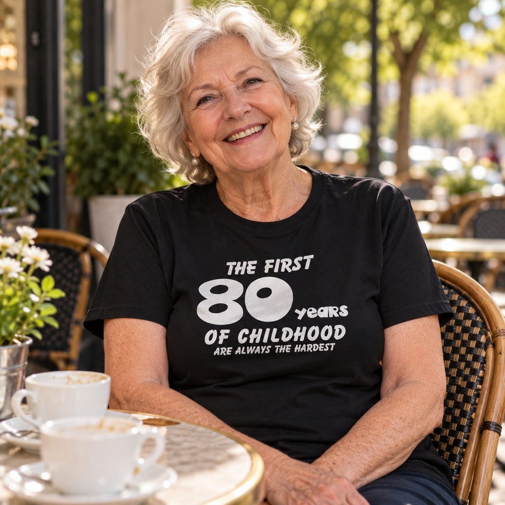 The First 80 Years of Childhood T-shirt in black - smiling woman at a café table with a cup of coffee, 80th birthday gift