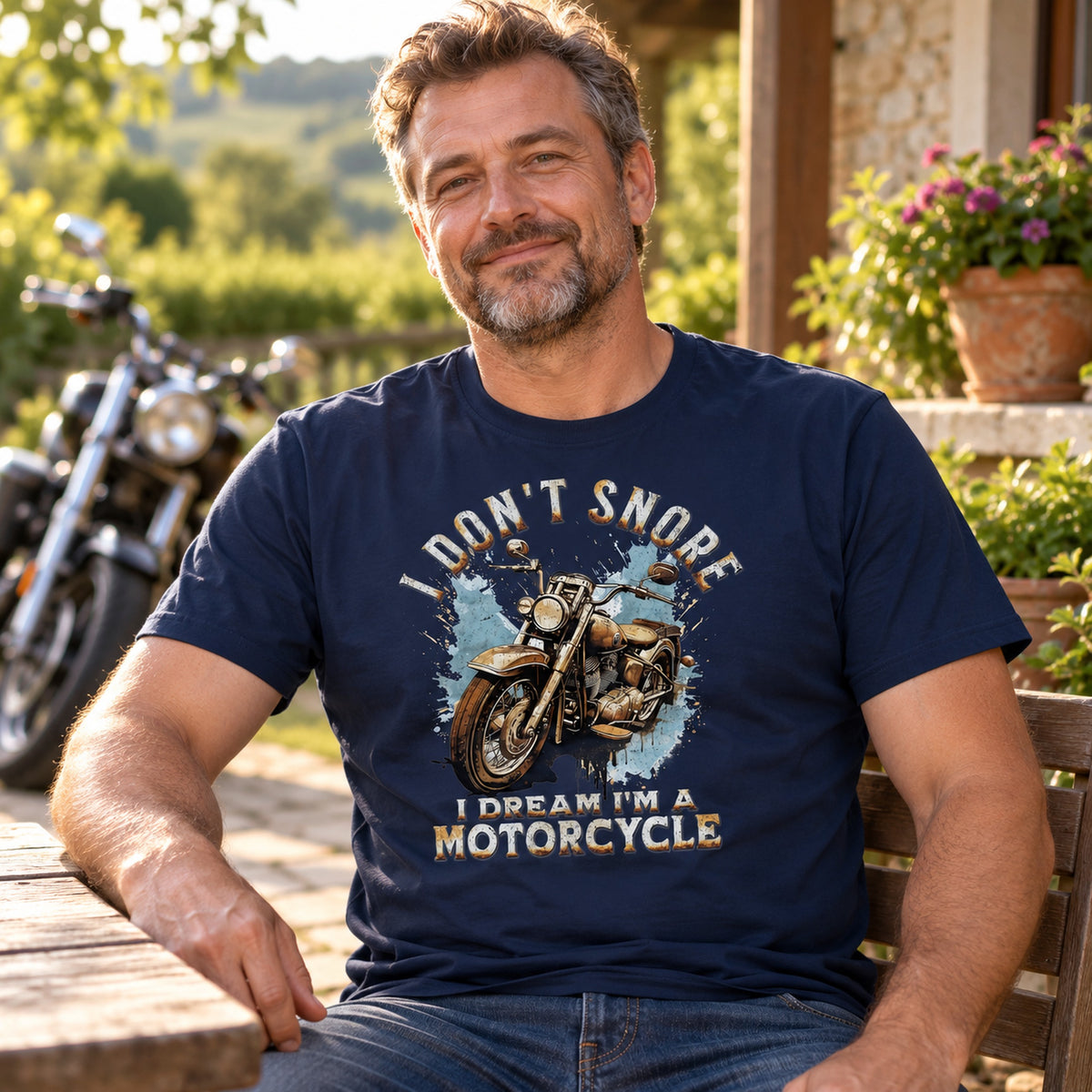 Biker in a navy I Don't Snore, I Dream I'm a Motorcycle t-shirt relaxing on a terrace, motorcycle in the background